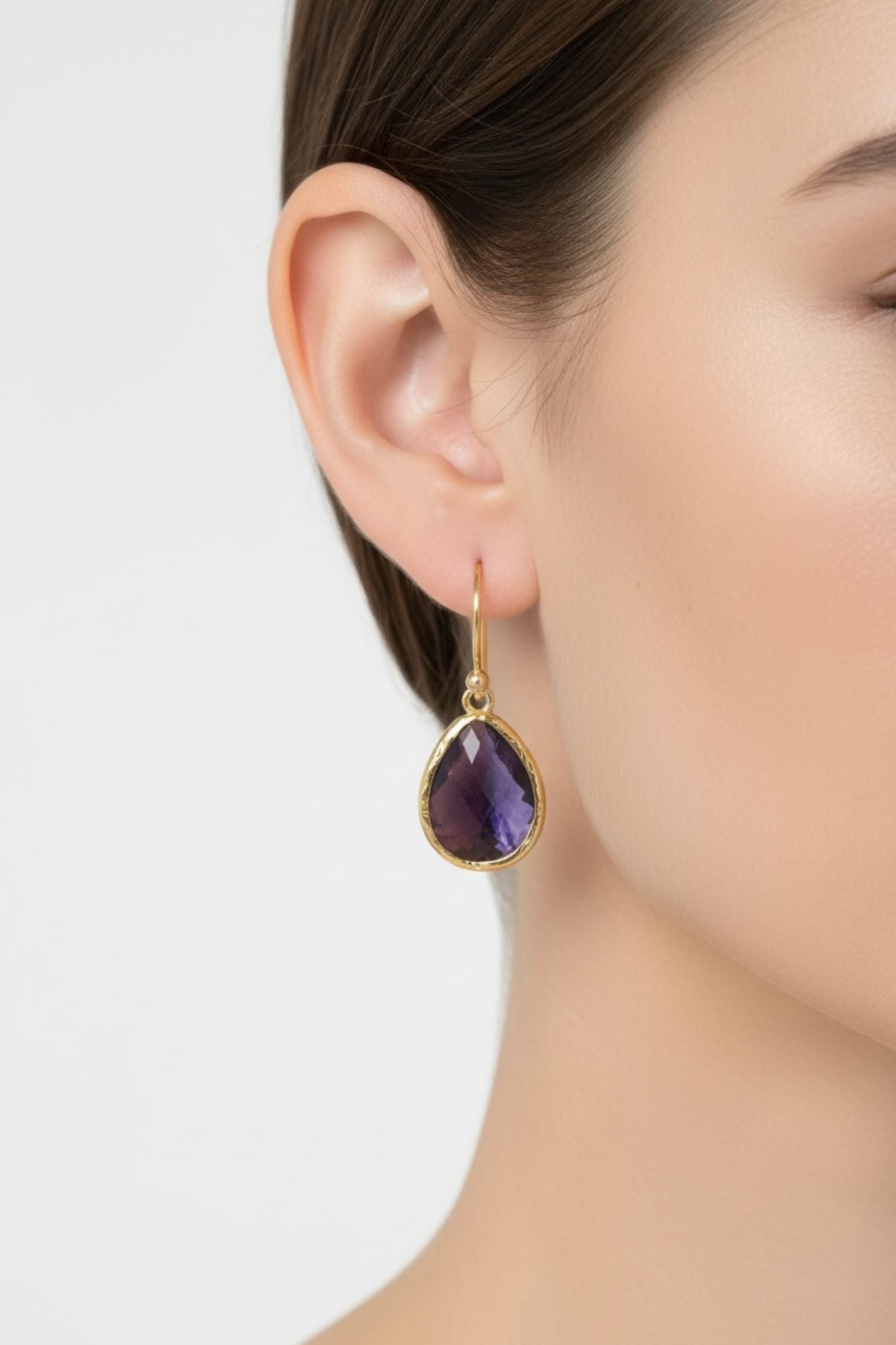 Close-up of a woman wearing a gold earring with a purple gemstone on a neutral background