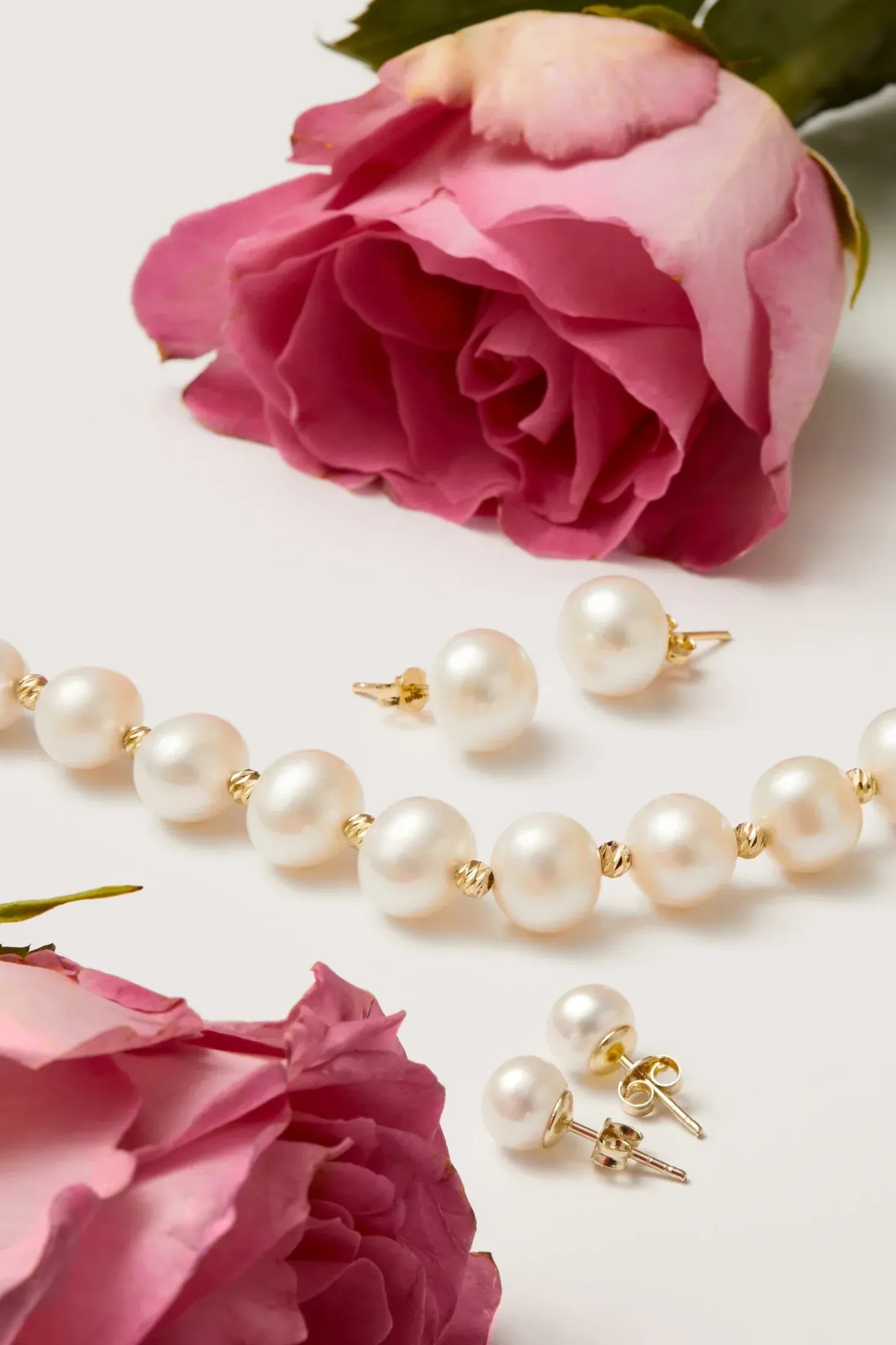 Pearl necklace and stud earrings alongside pink roses on a white surface.